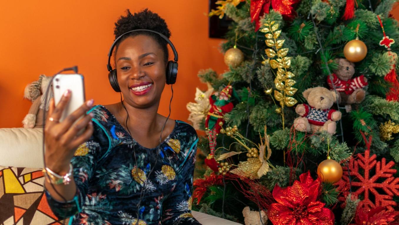 A photo of a woman of colour listening to content on a smartphone in front of a Christmas tree