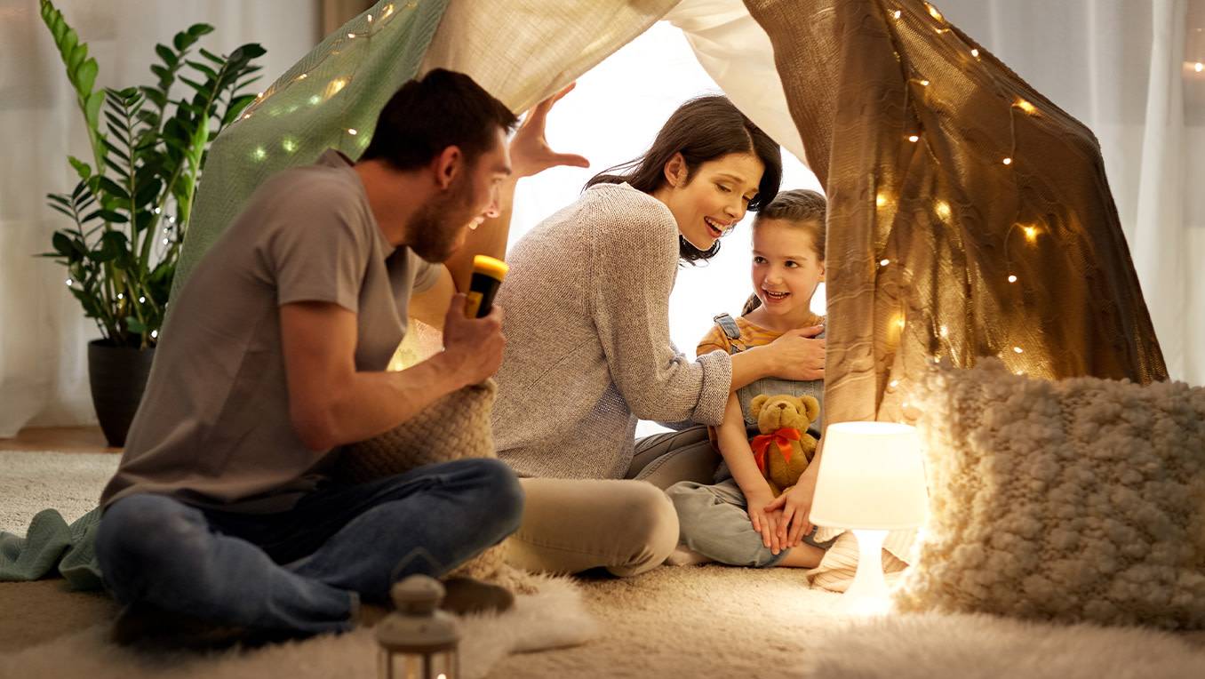 Father shares a story with mom and daughter in a homemade tent in their house with colourful lights around them