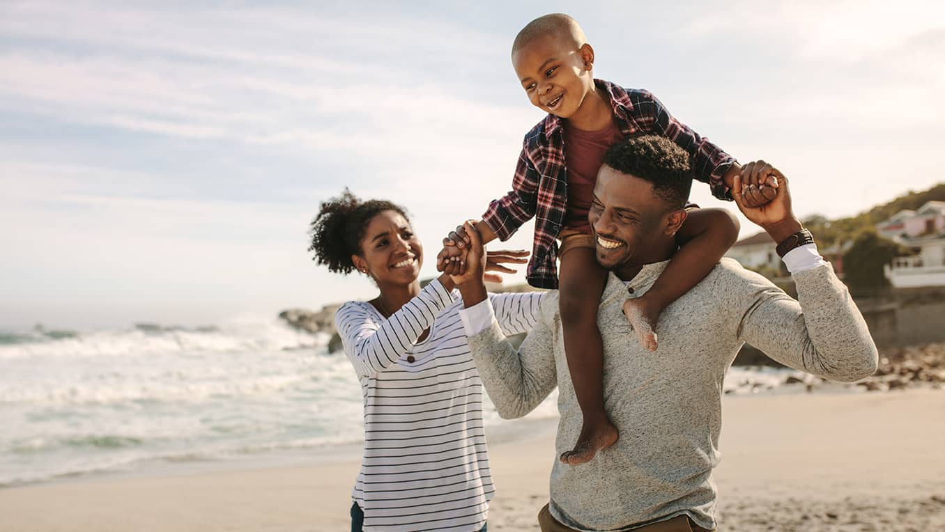 Happy parents carrying their smiling son on dad's shoulders while walking through the beach on a beautiful sunny day