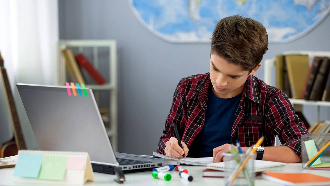 A teenage boy in a plaid shirt works on his homework at his desk with coloured markers, a notebook, and a laptop
