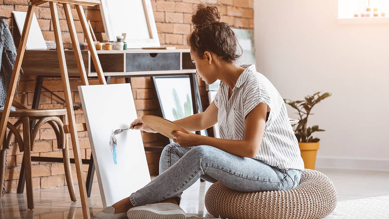 A young woman in jeans sits on the floor of a bright art studio and is putting the first brush strokes onto a fresh canvas