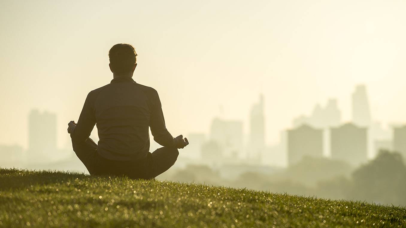 Silhouette of a man sitting in lotus position on grassy hilltop 