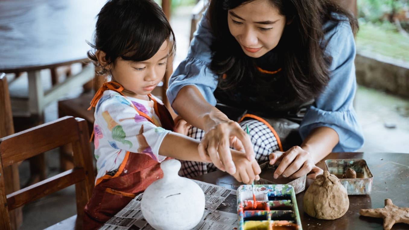 A mother and daughter paint pottery together at a wooden table