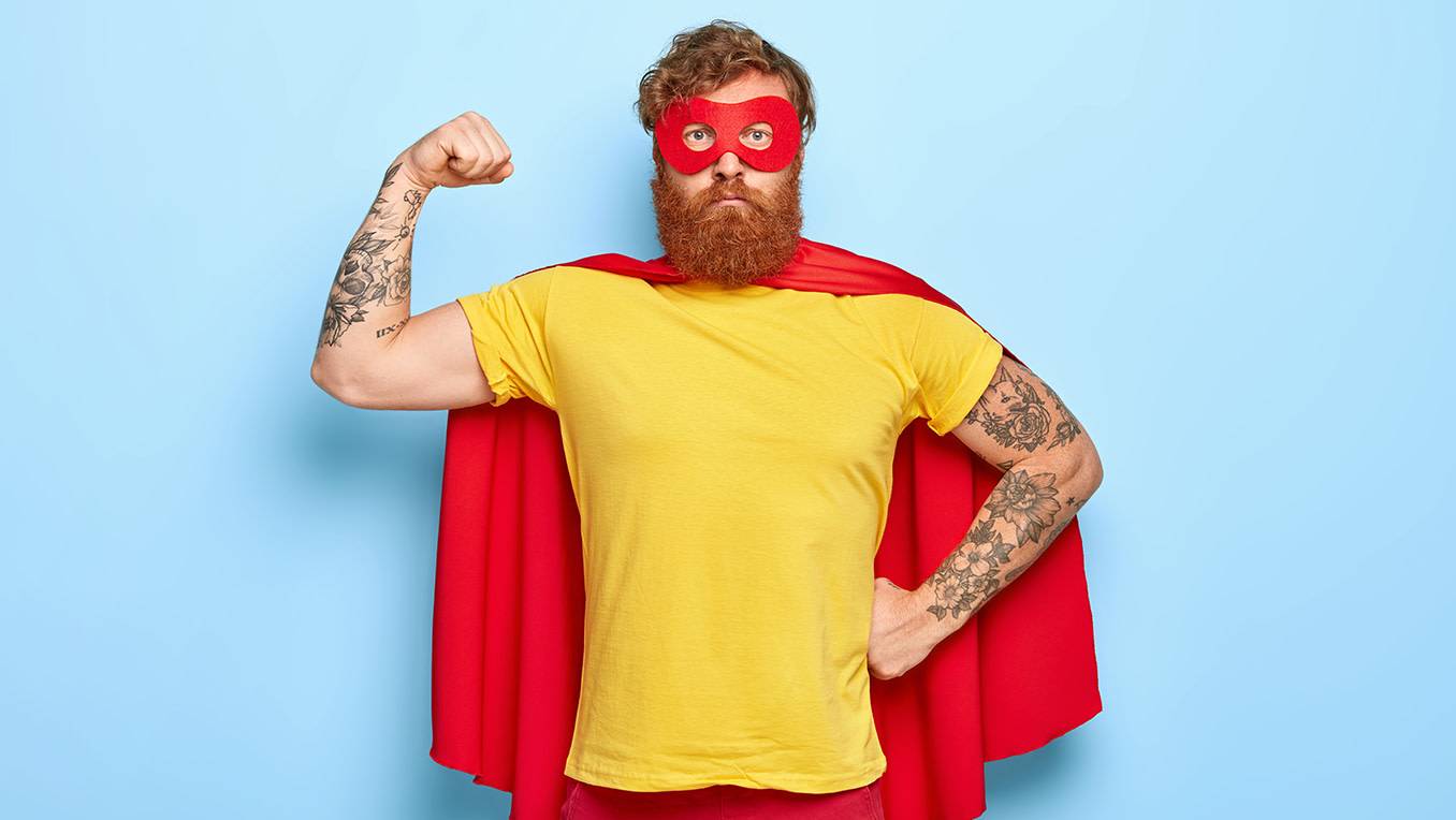 Bearded man wearing a red mask and red cape flexes his arm muscles in a super hero pose with a serious face