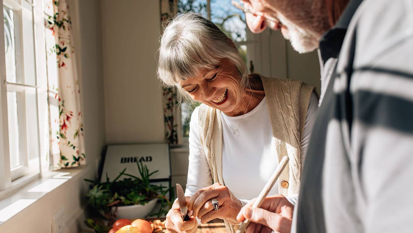 An elderly White couple peels vegetables in a sunlit kitchen, laughing