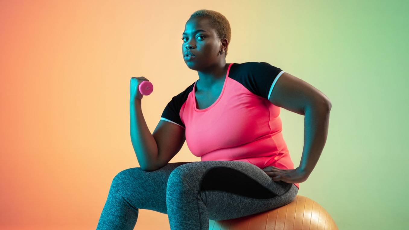 An image of a black woman sitting on a medicine ball lifting a dumbbell