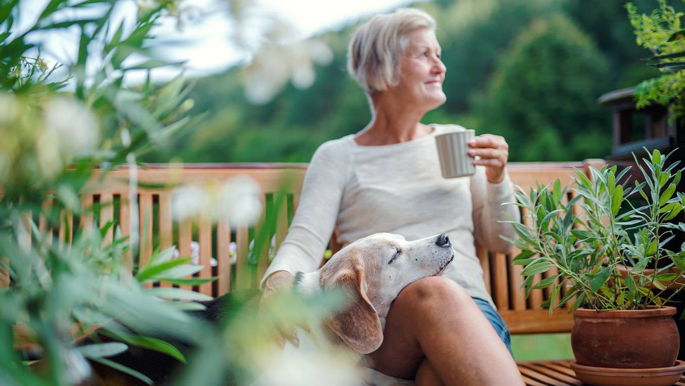 An elderly woman enjoys a cup of tea on a bench in a garden while her dog rests its head in her lap