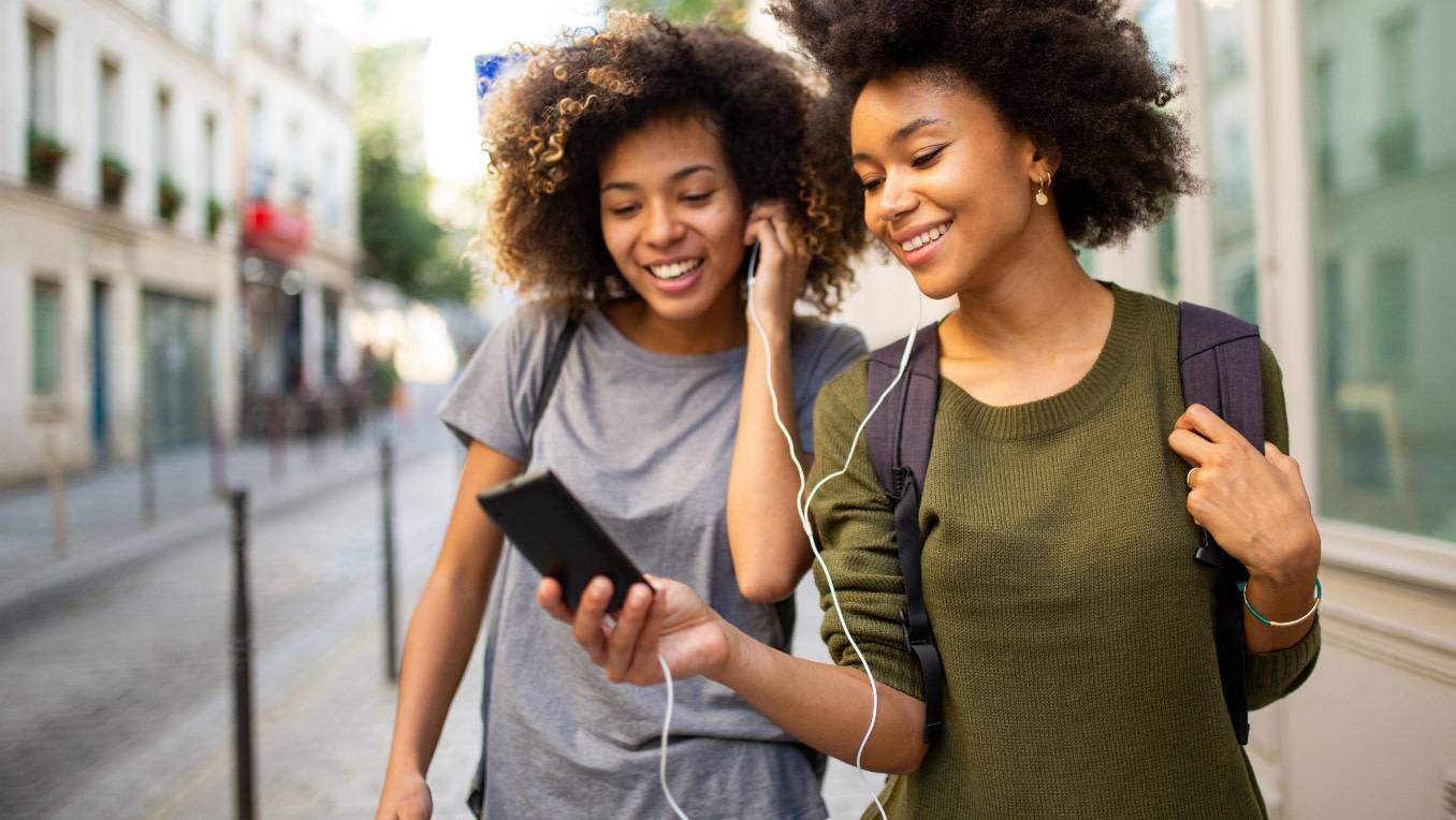 Two women happily share an audiobook as they walk down the street