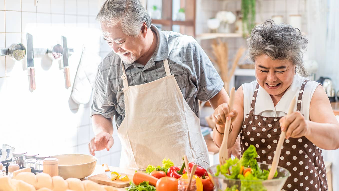 An elderly Asian couple prepare a meal together in a bright kitchen, laughing