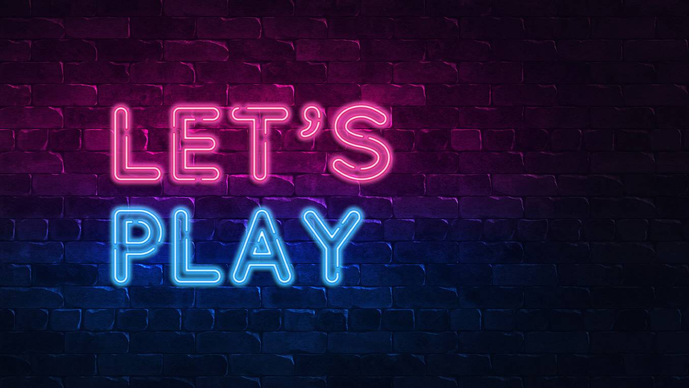 The words “Let’s Play” are lit in pink and blue neon against a darkened brick wall