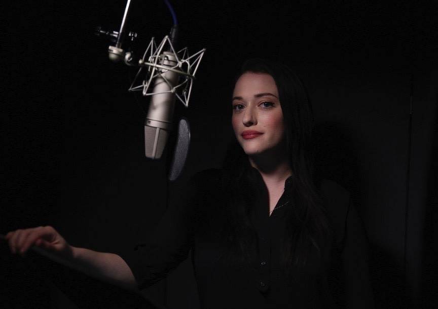 A photograph of actress Kat Dennings dressed in black, standing in a studio in front of a microphone