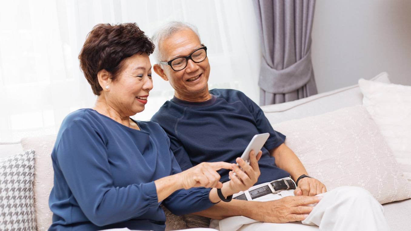 A senior Asian couple sitting together on a couch using a smartphone