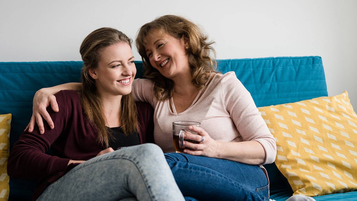 A mother with a cup of tea embraces her teenage daughter as they laugh together on a couch