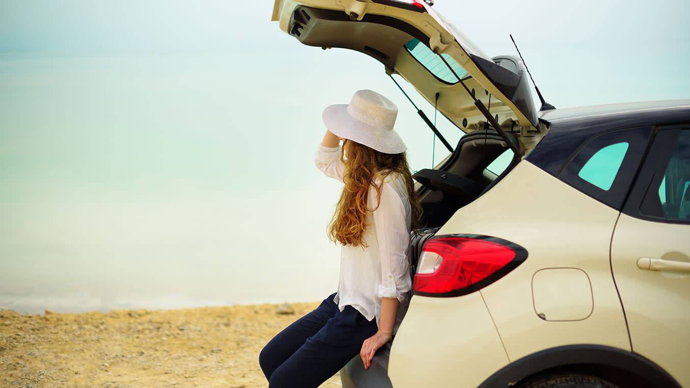 A young White woman in a beach hat sits on the open hatchback of her car on a beach and looks out to sea