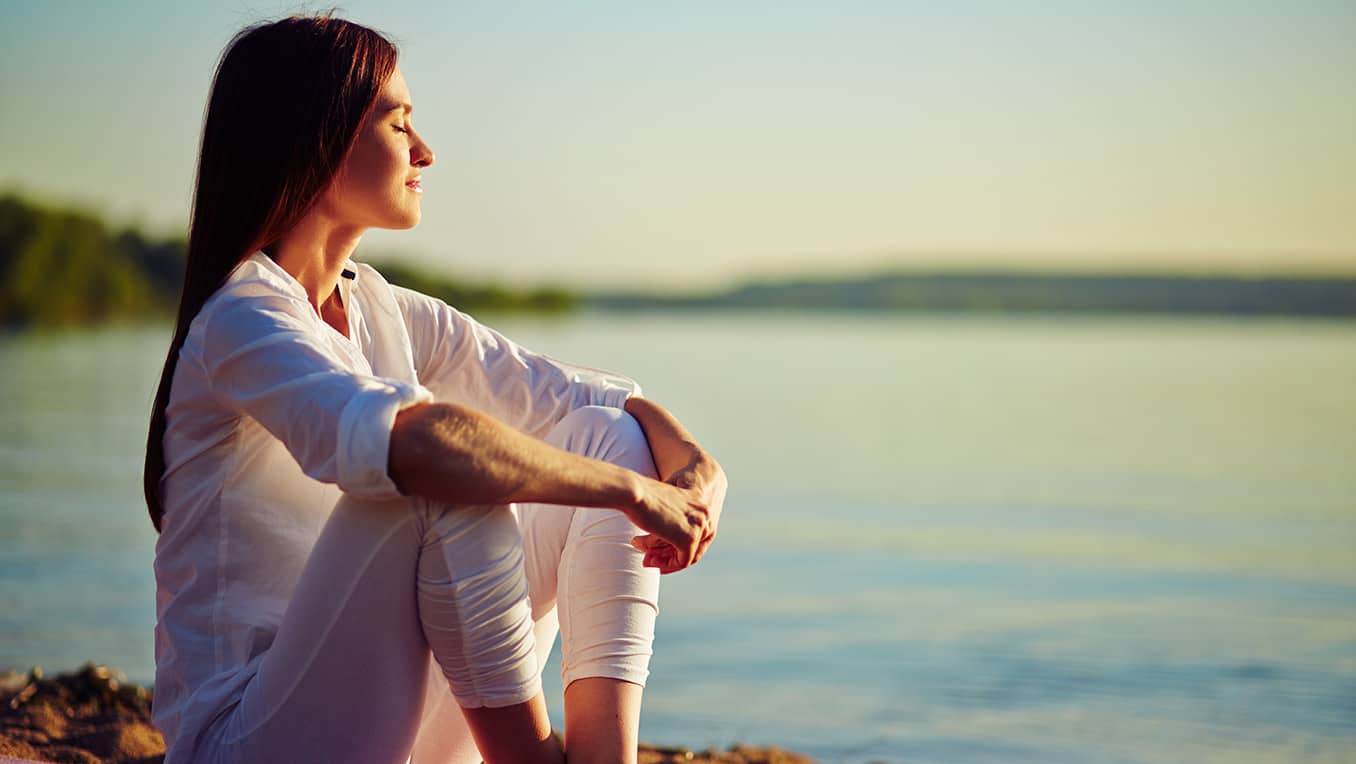 Femme paisiblement assise au soleil, face à l’eau, les yeux fermés