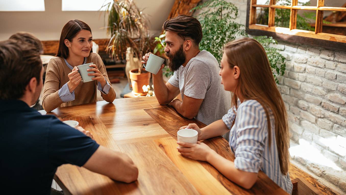 Group of two males and two females sit at a wooden coffee table drinking coffee having a group discussion