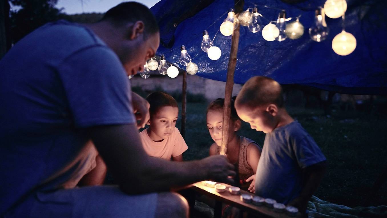 A father lighting candles with his young children as they camp outdoors under a canopy